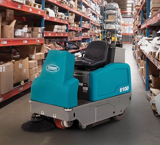 Tennant 6100 sub-compact battery rider sweeper working down a warehouse aisle between racking