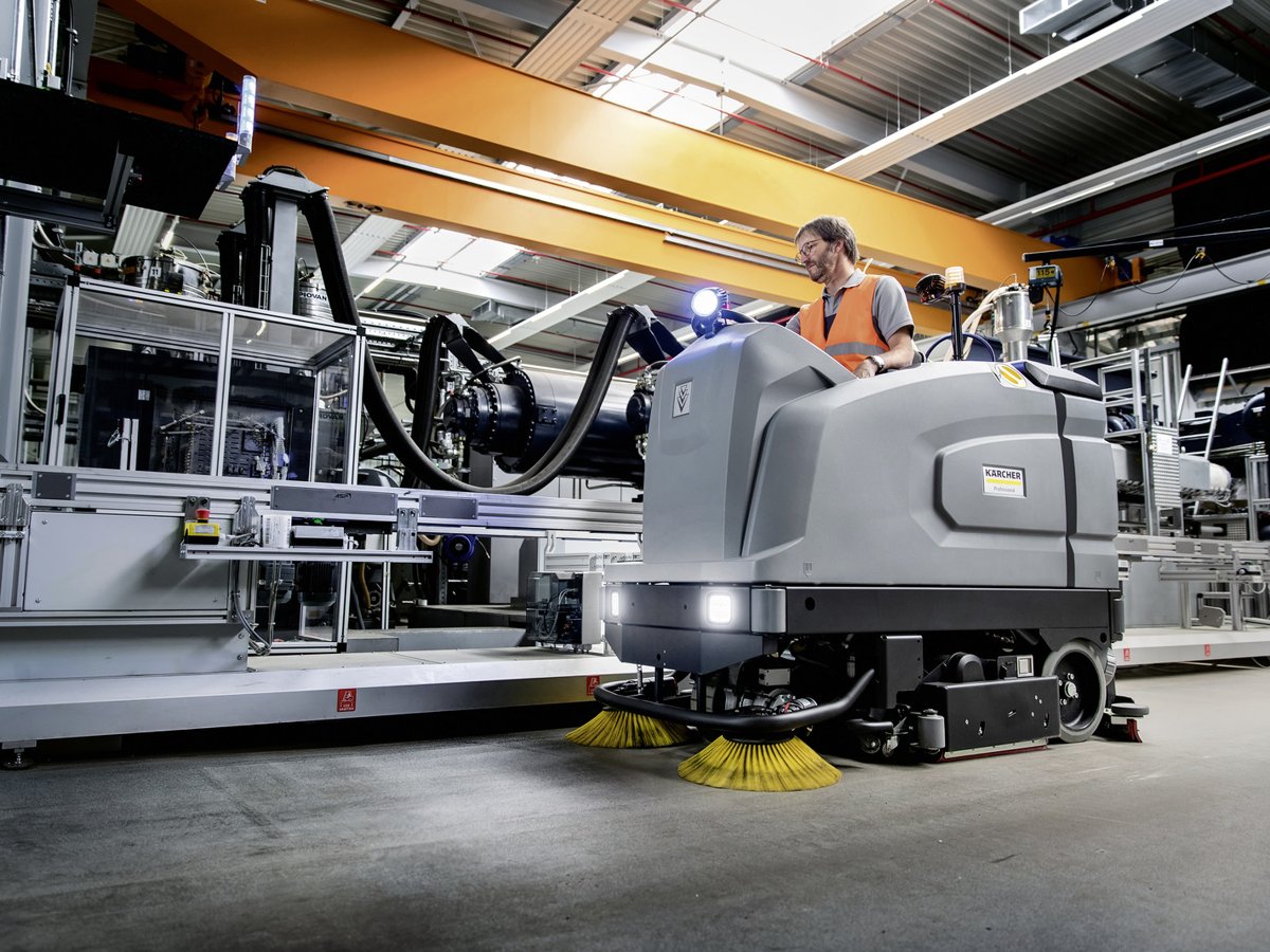 Operator riding a Kärcher B 260 RI scrubber through a factory aisle with lights on