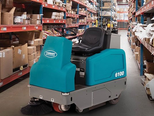 Tennant 6100 ride-on sweeper in a warehouse aisle between storage racks