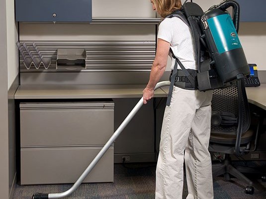 Operator vacuuming carpet under an office desk with a Tennant V-BP-6B